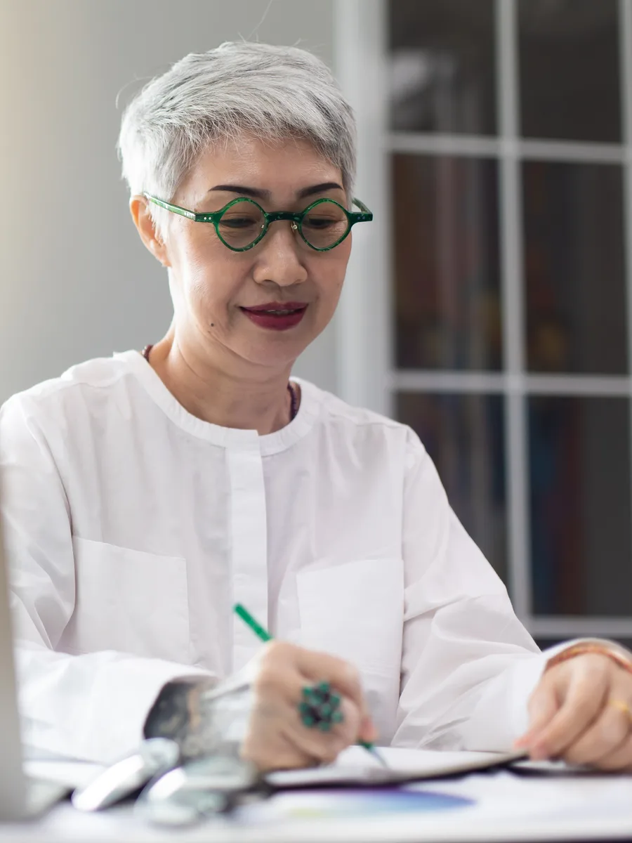 A woman in round green-rimmed glasses at a white desk, writing in an open notebook with a green pen, a bright window and bookshelves behind her.