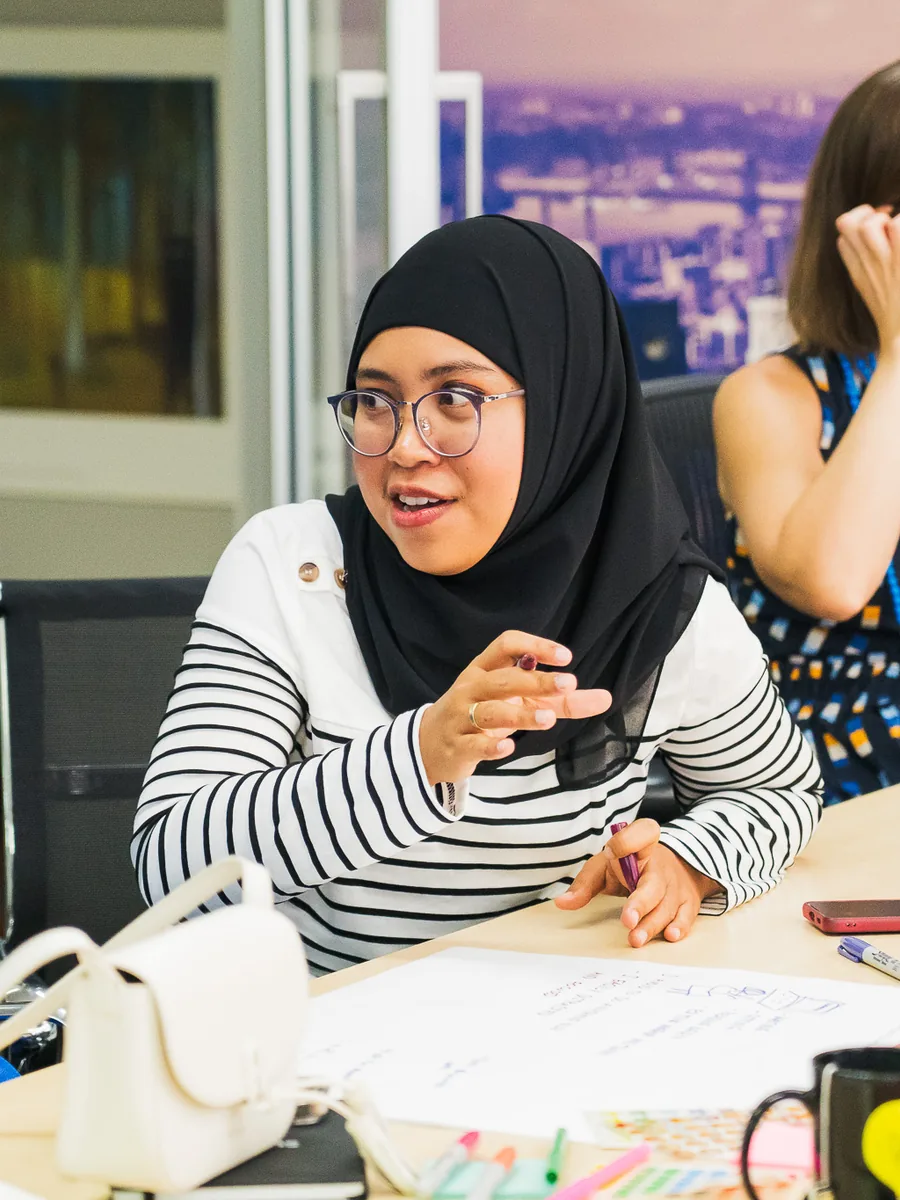 A woman in a hijab at a meeting table mid-sentence, hands raised, city windows behind her, papers and pens on the table.