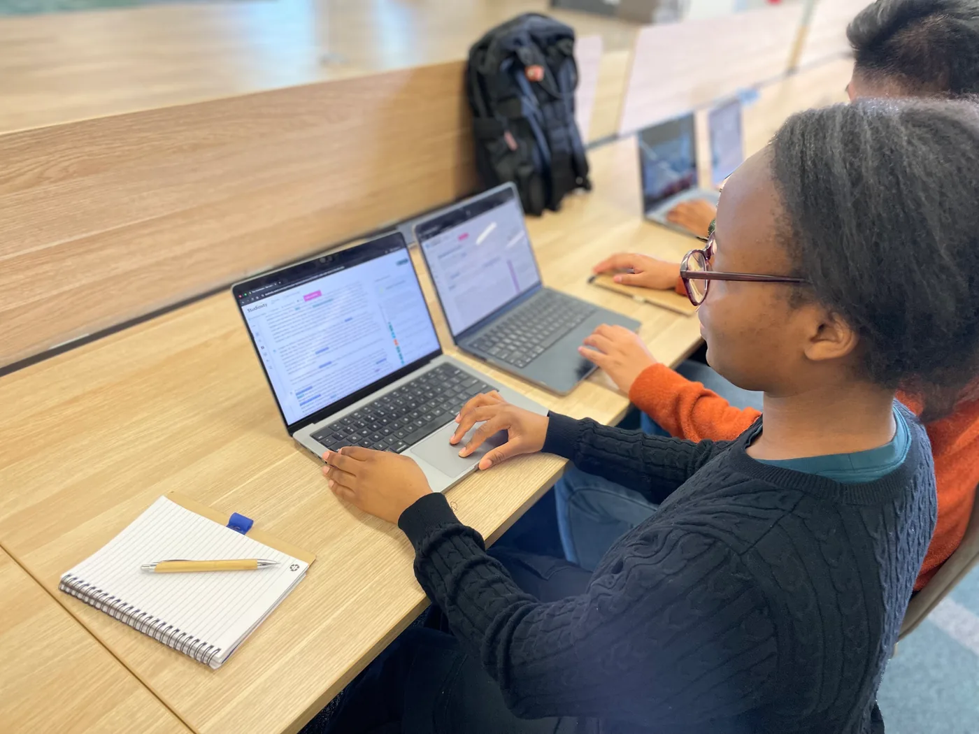 A student at a library desk with two laptops open, the nearer one showing a document with feedback listed beside it.