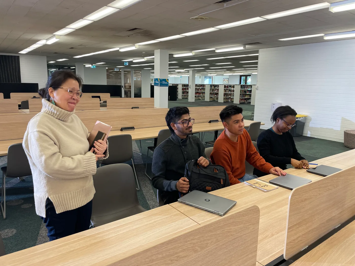 A university staff member standing beside three students seated at a library study bench, laptops and a notebook in front of them.