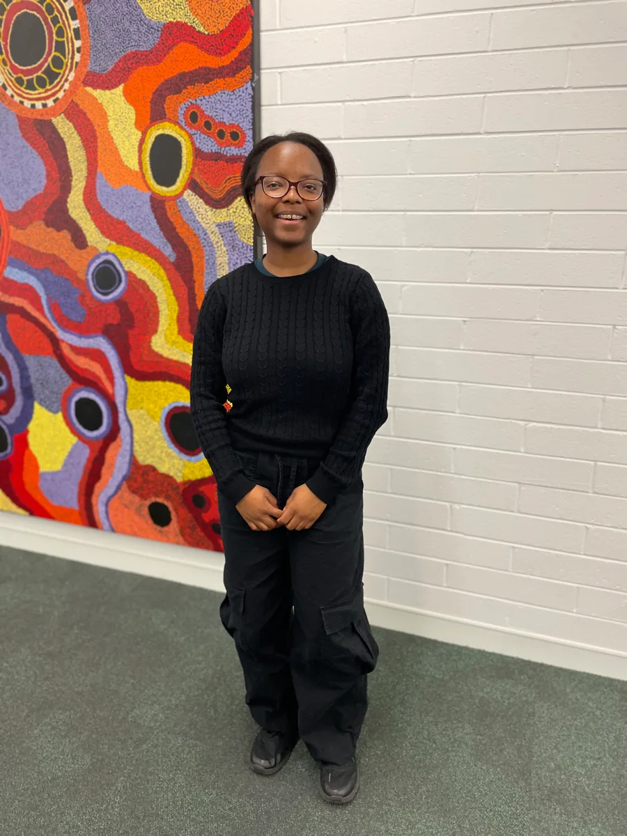 A student standing on campus in front of a large dot-painting mural beside a white brick wall.