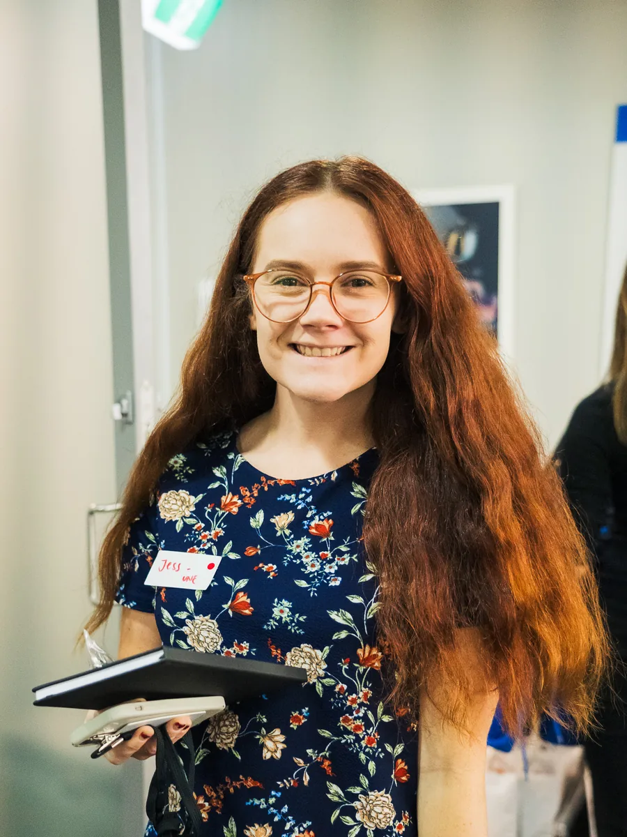 A woman in glasses standing facing camera in an office, notebooks and a pen held in front of her.