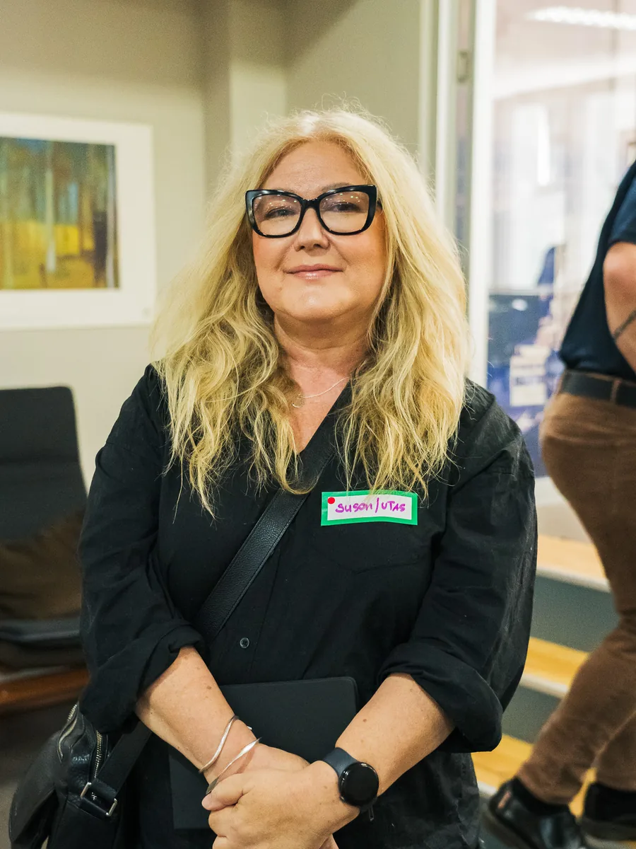 A woman in glasses standing facing camera in an office, a tablet held in front of her, a framed print on the wall behind.