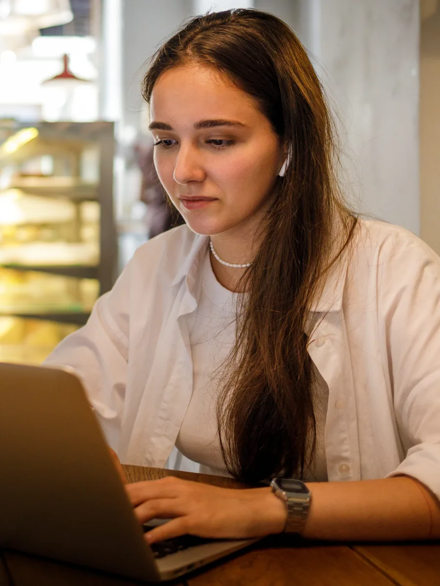 A student at a wooden café table typing on an open laptop, wearing an earphone, a lit display cabinet and the café interior behind her.