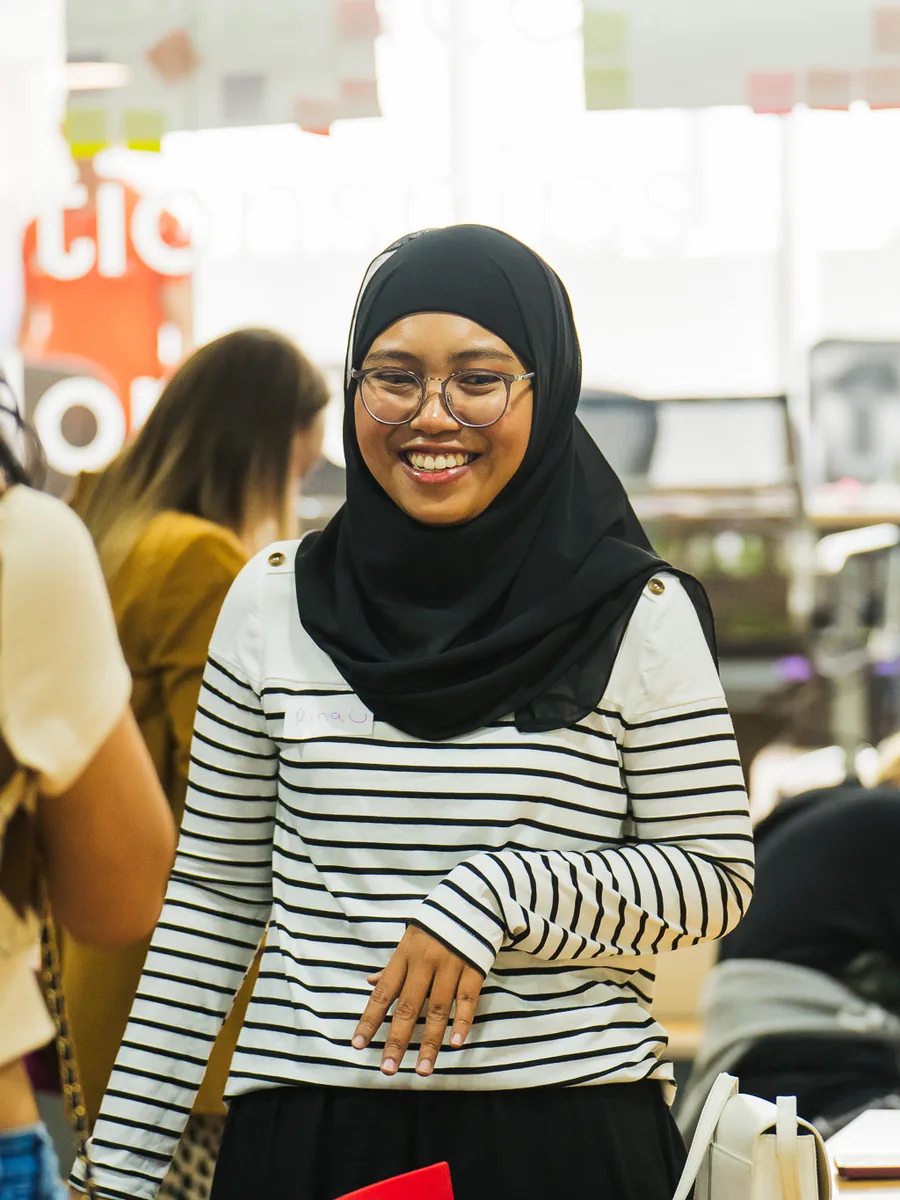 A student in a striped top smiling towards camera in a busy workshop room, a folder under her arm.