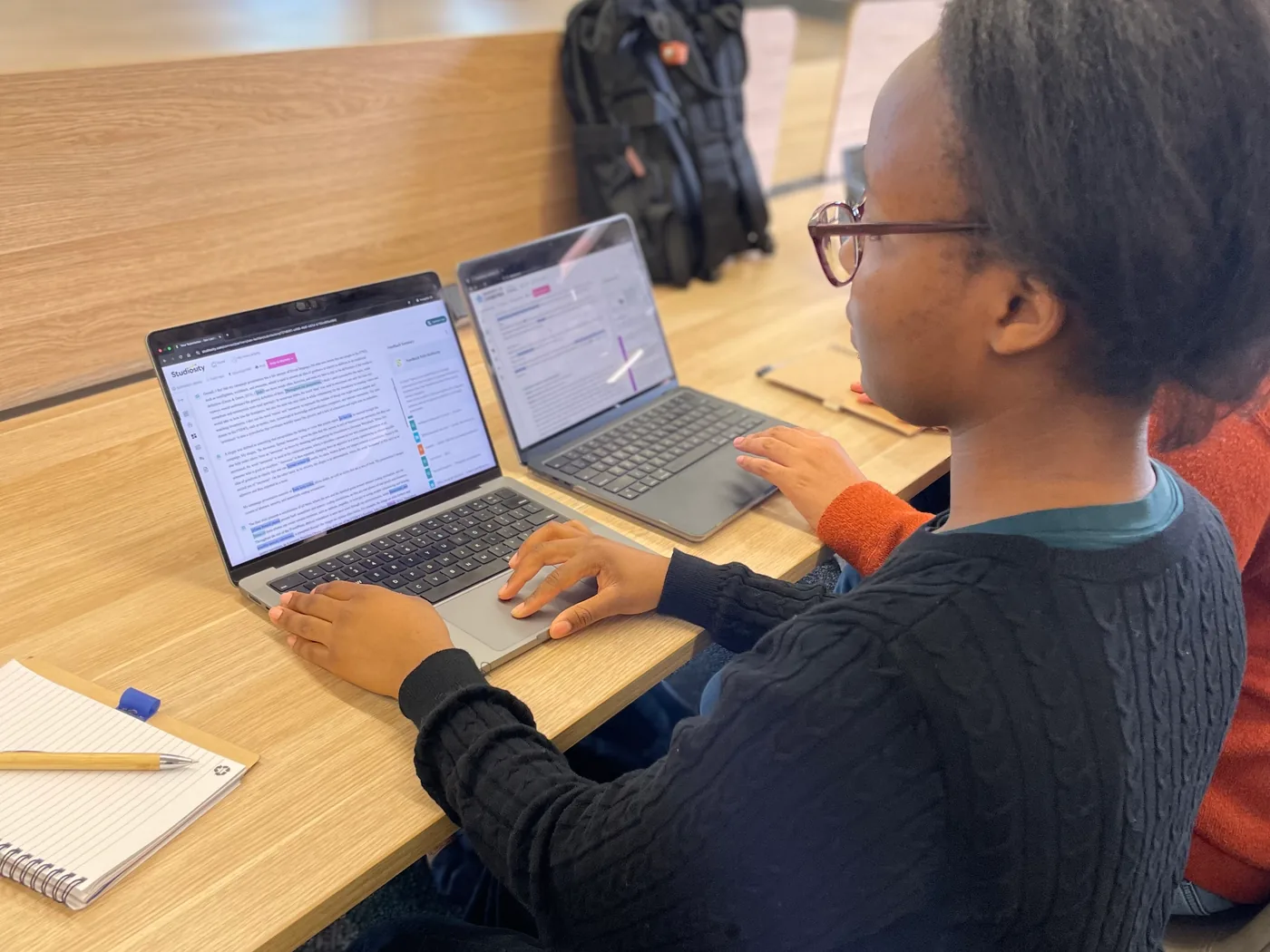 A student typing at a laptop in a library, a second laptop and an open notebook with a pen beside it.