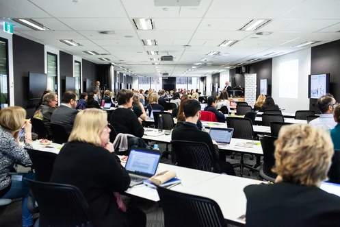 Delegates seated at long tables in a conference room, laptops open in front of them, a speaker presenting at the far end.